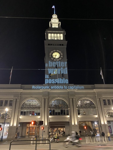 "off-white building with clock tower shows the following message projected onto it: a better world is possible, hashtag solarpunk: antidote to capitalism"