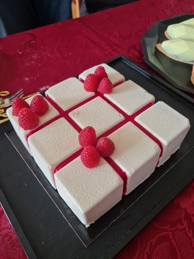 a white cocoa-butter sprayed mousse cake in the shape of the top layer of a rubik's cube with red seams and raspberries decoratively put on top